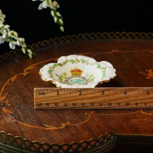 Op de afbeelding: Een kleine, witte, geschulpte schaal met gouden rand, met een kroon- en bloemenontwerp. De schaal heeft een diameter van ongeveer 7.6 cm. Een houten liniaal ligt naast de schaal.