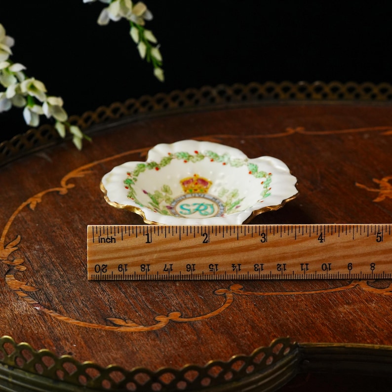 Op de afbeelding: Een kleine, witte, geschulpte schaal met gouden rand, met een kroon- en bloemenontwerp. De schaal heeft een diameter van ongeveer 7.6 cm. Een houten liniaal ligt naast de schaal.