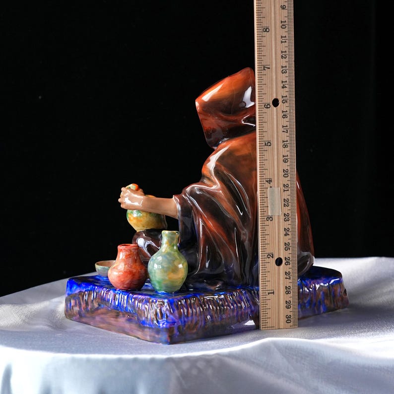 May include: A ceramic figurine of a robed figure, approximately 23 cm tall, seated and holding a small vessel. The figure is brown with a hood. Several small, colourful vases and bowls are in front of the figure. The base is blue and purple.