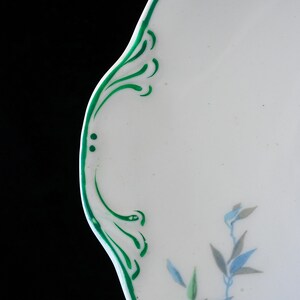 May include: White ceramic plate with a scalloped edge, adorned with green painted flourishes and delicate floral accents in blue and gray. The plate's design is a blend of classic and contemporary styles.
