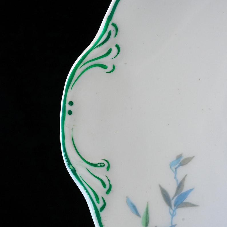 May include: White ceramic plate with a scalloped edge, adorned with green painted flourishes and delicate floral accents in blue and gray. The plate's design is a blend of classic and contemporary styles.