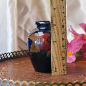 May include: A small, dark blue ceramic vase featuring a hand-painted floral design with red and orange petals and green leaves. A wooden ruler is placed vertically against the side of the vase, indicating a height of approximately 3.5 inches.