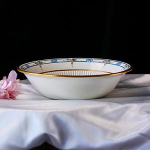 May include: White ceramic bowl with a gold rim and a decorative blue and floral border. The interior features a ribbed design. A pink flower is visible in the background. The bowl is likely vintage or antique.