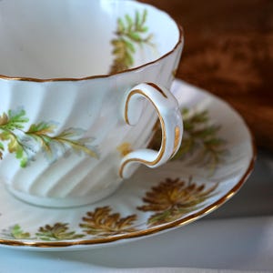 Op de afbeelding: Een witte theekop en schotel set met gouden randen en bloemmotieven in groen en goud. De theekop heeft een gebogen oor en een geribbelde buitenkant. De schotel heeft een bijpassend bloemmotief.
