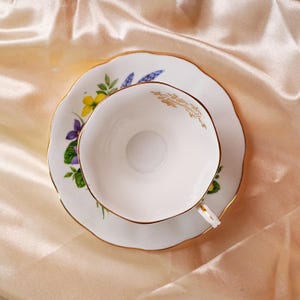 K&ouml;nnte beinhalten: Wei&szlig;es Tee-Set, bestehend aus Tasse und Untertasse mit floralem Muster in Gelb, Lila und Blau. Die R&auml;nder von Tasse und Untertasse sind goldfarben. Auf einer pfirsichfarbenen Satin-Oberfl&auml;che platziert.