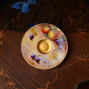 May include: A round porcelain saucer featuring a hand-painted design of colorful apples and grapes. The center of the saucer has a solid gold-toned circular indentation, and the rim is finished with a thin gold-colored border. The background is a soft, muted cream color with artistic brushstrokes.