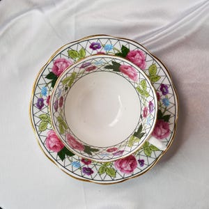 Op de afbeelding: Witte theekop en schotel set met een bloemenpatroon. De set heeft roze rozen, groene bladeren en kleine paarse en blauwe bloemen. De randen zijn goudkleurig afgewerkt. De theekop staat in het midden van de schotel.