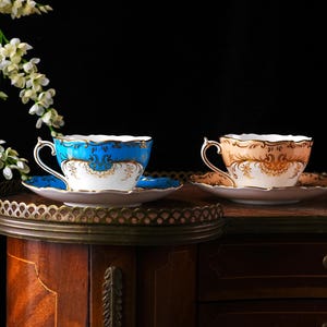 Op de afbeelding: Twee sierlijke theekopjes en schotels. E&eacute;n is blauw met gouden accenten, de andere perzik met gouden accenten. Beide hebben witte en gouden details. Ze staan op een decoratief dienblad op een houten kast, met witte bloemen op de achtergrond.