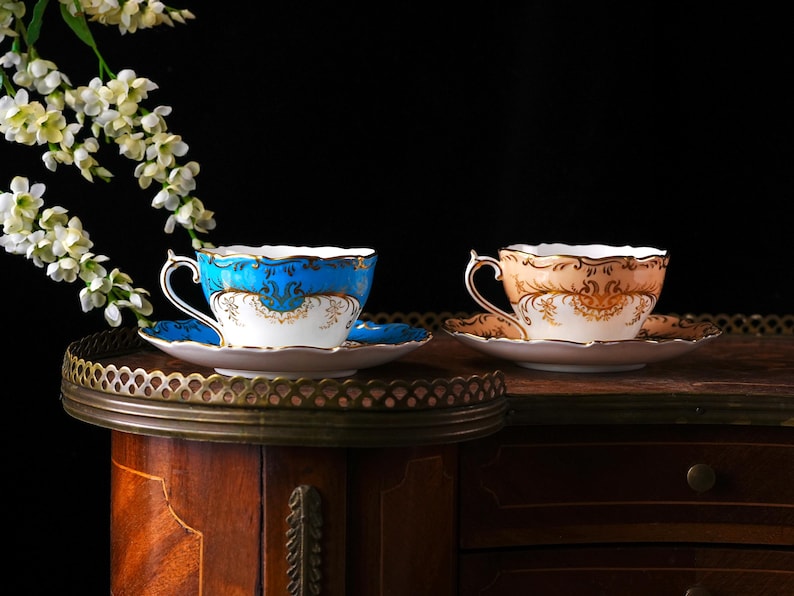 Op de afbeelding: Twee sierlijke theekopjes en schotels. E&eacute;n is blauw met gouden accenten, de andere perzik met gouden accenten. Beide hebben witte en gouden details. Ze staan op een decoratief dienblad op een houten kast, met witte bloemen op de achtergrond.