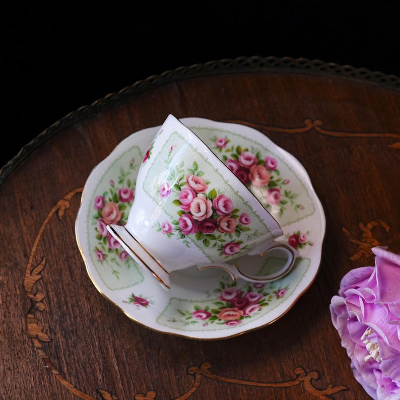 K&ouml;nnte beinhalten: Ein Vintage-Tee-Set, bestehend aus einer Tasse und einer Untertasse. Die wei&szlig;e Porzellantasse und Untertasse zeigen ein zartes Blumenmuster aus rosa Rosen und gr&uuml;nem Laub auf hellgr&uuml;nem Hintergrund. Goldene Verzierungen akzentuieren die R&auml;nder. Das Set steht auf einer dunklen Holzoberfl&auml;che.