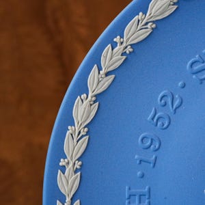 May include: Close-up of a blue ceramic plate with a white floral border. The plate has embossed white leaves and small flower details. The plate has the embossed text "H.1.9.52."