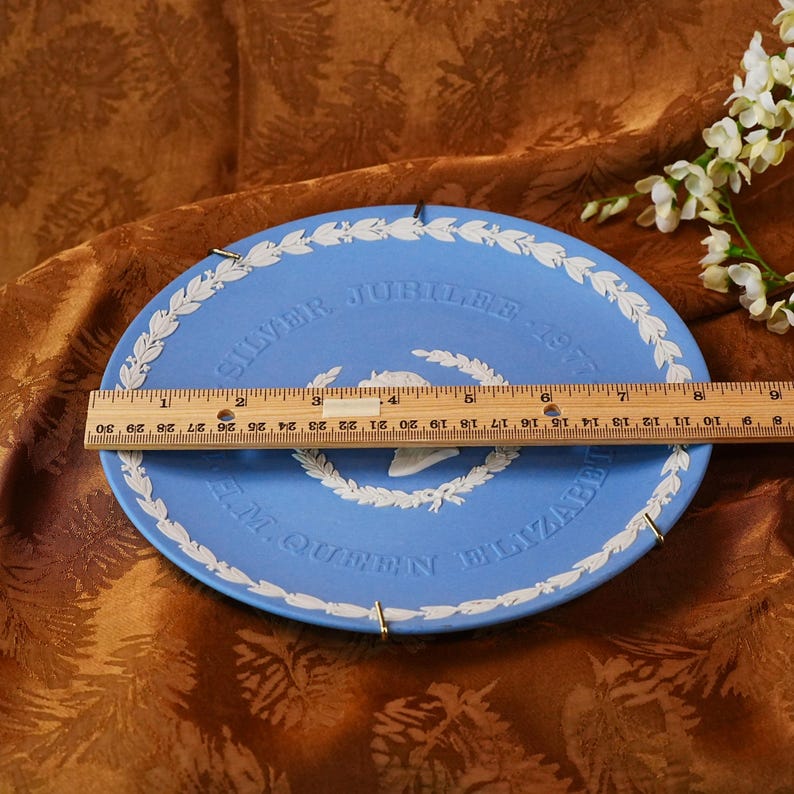 May include: A light blue commemorative plate with white lettering and a white laurel wreath design. The plate reads "SILVER JUBILEE 1977" and "H.M. QUEEN ELIZABETH". The plate measures approximately 25 cm in diameter.