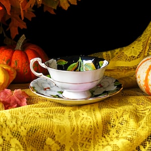 May include: A white teacup and saucer set with a black interior featuring green and yellow leaf designs. The saucer and cup exterior have white floral patterns. The set is placed on a yellow lace tablecloth, with pumpkins and fall leaves in the background.