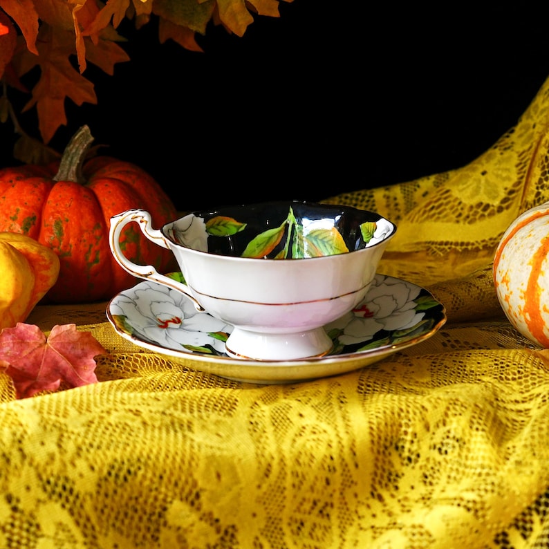 May include: A white teacup and saucer set with a black interior featuring green and yellow leaf designs. The saucer and cup exterior have white floral patterns. The set is placed on a yellow lace tablecloth, with pumpkins and fall leaves in the background.