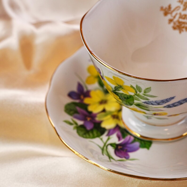 K&ouml;nnte beinhalten: Eine wei&szlig;e Teetasse und Untertasse mit einem Blumendesign aus gelben und lila Blumen, gr&uuml;nen Bl&auml;ttern und goldenen Verzierungen. Das Set befindet sich vor einem cremefarbenen Hintergrund.
