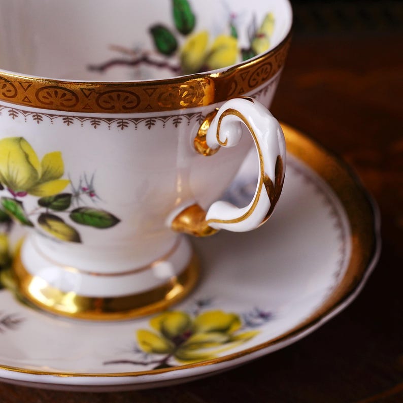 Peut inclure: Une tasse et une soucoupe blanches avec un bord et une anse dor&eacute;s. La tasse et la soucoupe sont d&eacute;cor&eacute;es de motifs floraux jaunes et d'accents dor&eacute;s. La tasse mesure environ 7.6 cm de haut.