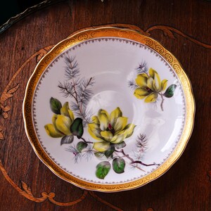 Peut inclure: Une assiette en porcelaine blanche avec un bord dor&eacute; et un motif floral. L'assiette pr&eacute;sente des fleurs de magnolia jaunes avec des feuilles vertes et des aiguilles de pin grises. Le bord a un motif dor&eacute; d&eacute;coratif. L'assiette est sur une surface en bois fonc&eacute;.