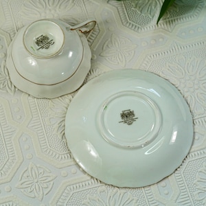 May include: White teacup and saucer set with gold trim. The teacup is upside down, revealing a logo. The saucer is round with a scalloped edge. The set is on a white, textured surface.