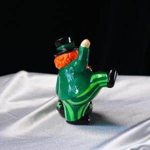 May include: A ceramic figurine of a leprechaun in a green suit with a black top hat. The suit has a striped pattern on the pants and a textured jacket. The leprechaun has red hair and is posed with one leg raised.
