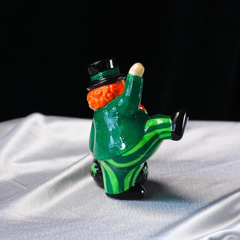 May include: A ceramic figurine of a leprechaun in a green suit with a black top hat. The suit has a striped pattern on the pants and a textured jacket. The leprechaun has red hair and is posed with one leg raised.