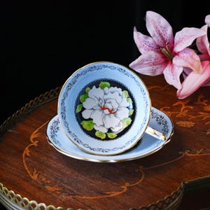 May include: A light blue teacup and saucer set featuring a central white peony illustration with green leaves on a black circular background. The rim of the cup and saucer is decorated with a delicate black filigree pattern and a thin gold trim.