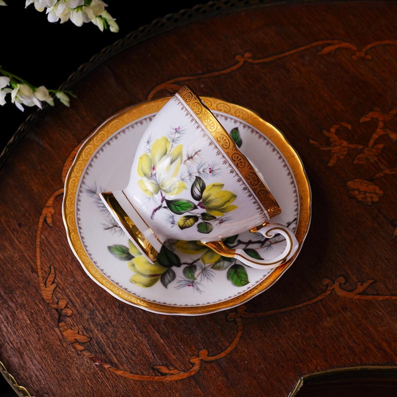Peut inclure: Un service &agrave; th&eacute; blanc avec une tasse et une soucoupe, orn&eacute; d'un motif floral jaune et de bordures dor&eacute;es. La tasse repose sur la soucoupe, &eacute;galement d&eacute;cor&eacute;e du m&ecirc;me motif floral et d'accents dor&eacute;s. Sur une surface en bois.