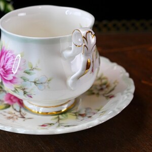 May include: A white teacup and saucer set with a floral design featuring pink and purple flowers, green leaves, and gold detailing. The teacup has a decorative handle and sits on a matching saucer.