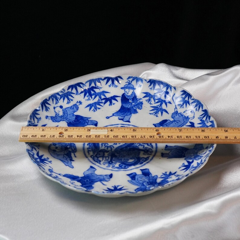 Puede incluir: Un plato de cer&aacute;mica blanco y azul con borde festoneado. El plato presenta un dise&ntilde;o pintado en azul de figuras y bamb&uacute;. Una regla muestra que el plato tiene aproximadamente 30 cm de di&aacute;metro.