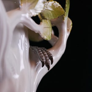 May include: A detailed view of a white porcelain sculpture featuring a bird's claw grasping a branch with green and yellow leaves. The sculpture has a glossy finish and is set against a black background.