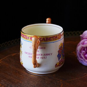 Puede incluir: Taza de cer&aacute;mica blanca con borde dorado y asa en forma de pierna. La taza presenta texto e ilustraciones coloridas, incluyendo las palabras "Queen Elizabeth" y "Westminster Abbey May 12 1937".