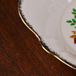 May include: A white ceramic plate with a scalloped edge featuring a gold-trimmed rim and a black speckled border. The plate displays a partial graphic of green leaves and yellow wheat stalks tied with a red ribbon on its surface.