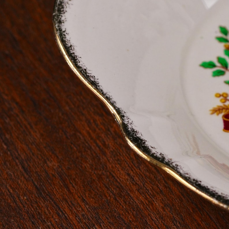 May include: A white ceramic plate with a scalloped edge featuring a gold-trimmed rim and a black speckled border. The plate displays a partial graphic of green leaves and yellow wheat stalks tied with a red ribbon on its surface.