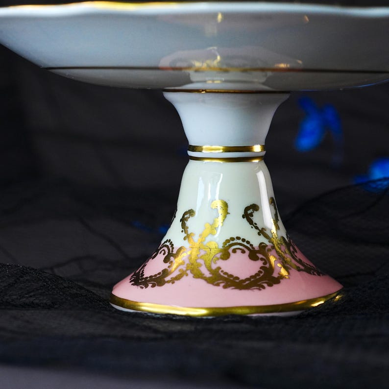 Puede incluir: Un soporte para pasteles de porcelana blanca con borde festoneado y ribete dorado. La base est&aacute; decorada con una banda rosa suave y patrones intrincados de filigrana dorada, acentuados por bandas doradas horizontales en el tallo central.