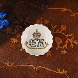 Op de afbeelding: Een kleine, ronde, wit keramische schaal met een geschulpte rand, voorzien van een centrale kroonafbeelding boven de letters E en A. Een rood lint onderaan bevat de tekst: EDWARDUS VII D.G. BRITT OMN. REX F.D. IND. IMP. CROWNED 26 JUNE 1902.