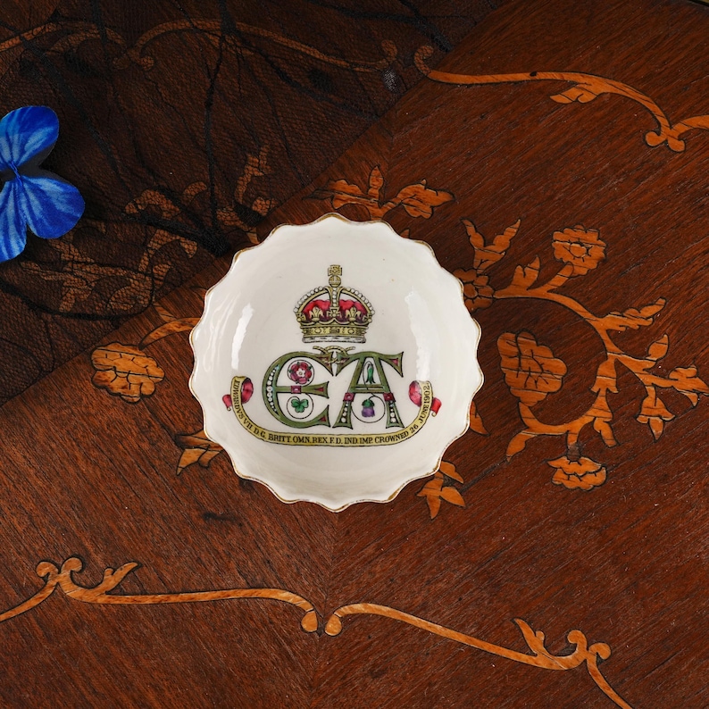 Op de afbeelding: Een kleine, ronde, wit keramische schaal met een geschulpte rand, voorzien van een centrale kroonafbeelding boven de letters E en A. Een rood lint onderaan bevat de tekst: EDWARDUS VII D.G. BRITT OMN. REX F.D. IND. IMP. CROWNED 26 JUNE 1902.