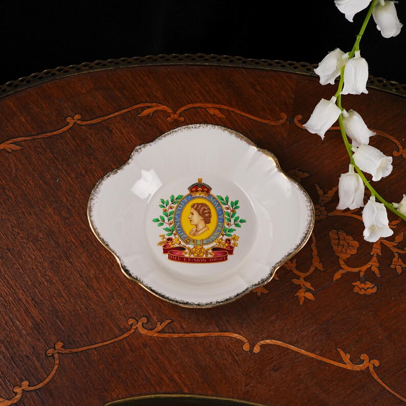 May include: A white porcelain dish with a scalloped edge and gold trim. The center features a colorful royal emblem with a profile portrait of Queen Elizabeth II, a crown, and the text Dieu et mon droit on a red ribbon banner.