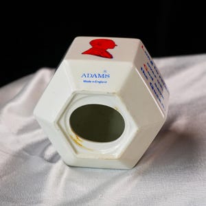 Op de afbeelding: Witte keramische spaarpot met een geometrisch ontwerp. Voorzien van een rood silhouet en de tekst "God save the Queen". De spaarpot heeft een gleuf voor munten en de merknaam "ADAMS Made in England".