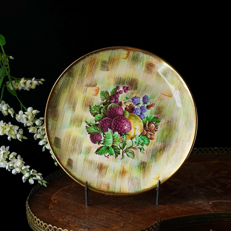 May include: A vintage decorative plate with a gold rim and a painted fruit design. The plate features a central arrangement of purple berries, green leaves, and a yellow fruit against a textured, light green and gold background.