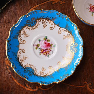 Op de afbeelding: Een vintage theekopschotel met een helderblauwe rand en gouden details. Het witte midden heeft een roze rozenbloemmotief. De schotel ligt op een donker houten oppervlak.