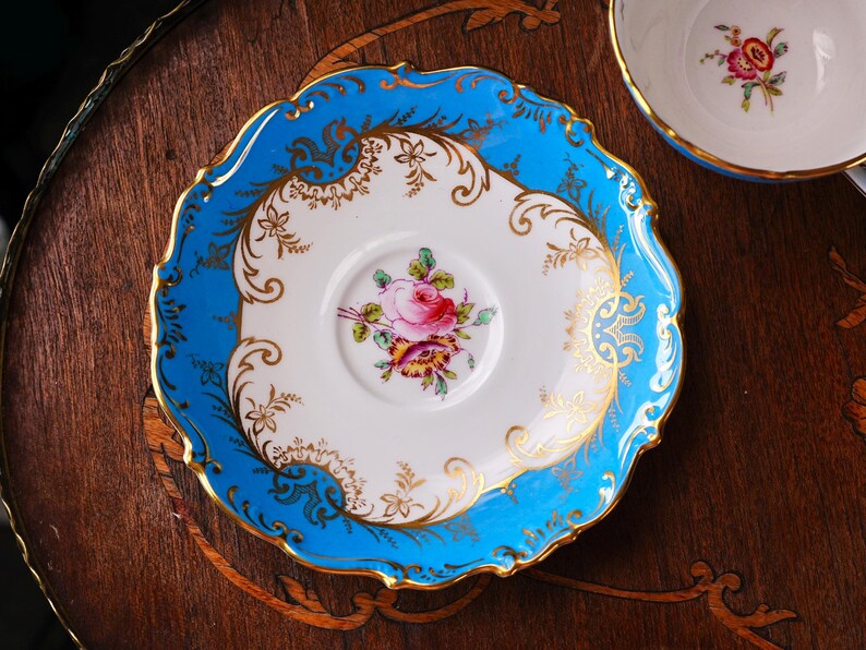 Op de afbeelding: Een vintage theekopschotel met een helderblauwe rand en gouden details. Het witte midden heeft een roze rozenbloemmotief. De schotel ligt op een donker houten oppervlak.