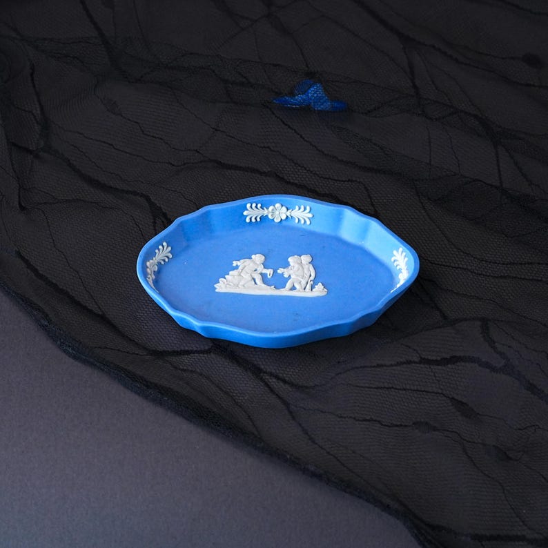 May include: An oval blue ceramic tray featuring white relief figures of two people interacting in the centre, with white floral and leaf-like decorative accents along the scalloped rim, showcasing a classic matte finish and raised detailing.