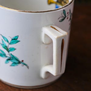 May include: White ceramic mug with a gold rim and handle accents. The mug features hand-painted teal leaves and a gold design near the rim. The handle is square and has a gold stripe down the center.