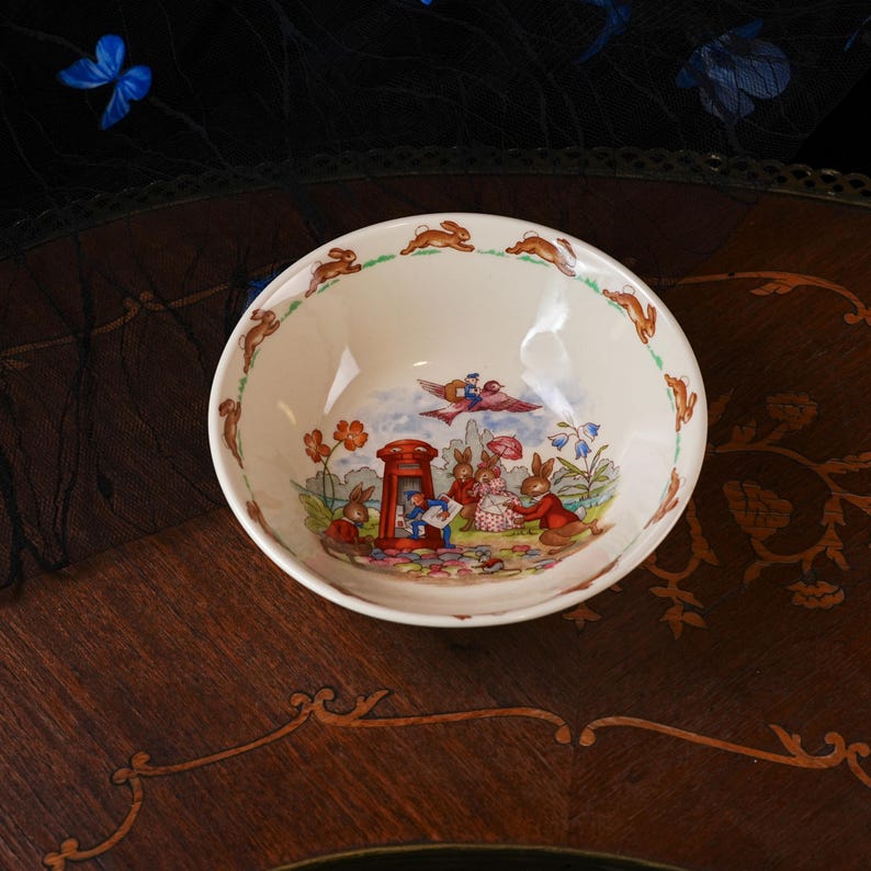 May include: A cream-colored ceramic bowl featuring a repeating border of brown rabbits around the rim. The center of the bowl displays a colorful illustration of anthropomorphic rabbits near a red pillar box, with flowers and a bird overhead.