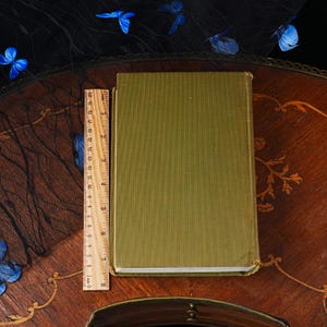 Puede incluir: Un libro de tapa dura rectangular con una cubierta de tela verde oliva texturizada. Una regla de madera est&aacute; colocada verticalmente a lo largo del lado izquierdo del libro, mostrando una longitud de unos 20 cm para la cubierta.