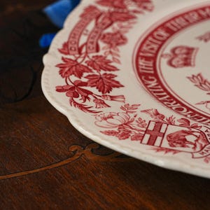 May include: A cream-coloured ceramic plate with a scalloped edge, featuring a red transferware pattern. The design includes floral motifs, a crown, and text reading COMMEMORATING THE VISIT OF THEIR MAJESTIES and DOMINION OF CANADA.