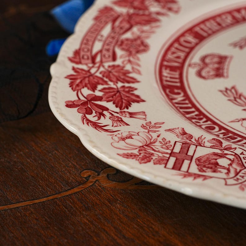May include: A cream-coloured ceramic plate with a scalloped edge, featuring a red transferware pattern. The design includes floral motifs, a crown, and text reading COMMEMORATING THE VISIT OF THEIR MAJESTIES and DOMINION OF CANADA.