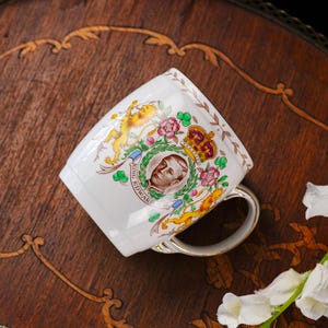 Może przedstawiać: Biały ceramiczny kubek z pamiątkowym portretem kr&oacute;la Edwarda VIII, otoczony kolorowym wzorem heraldycznym z koroną, lwem i motywami kwiatowymi. Napis King Edward VIII jest wydrukowany wewnątrz okrągłej ramki.