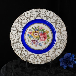 Puede incluir: Un plato de porcelana vintage blanco con un patrón de filigrana dorada. El plato presenta un vibrante ramo floral en el centro, rodeado por un borde azul brillante. El plato se exhibe sobre un soporte negro. Diámetro aproximado de 25 cm.