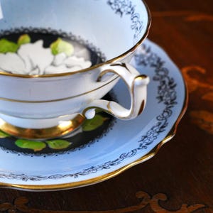 May include: A light blue porcelain teacup and saucer set featuring a white floral design with green leaves against a black circular background. The set is accented with a delicate black floral border pattern and finished with a gold rim and handle.