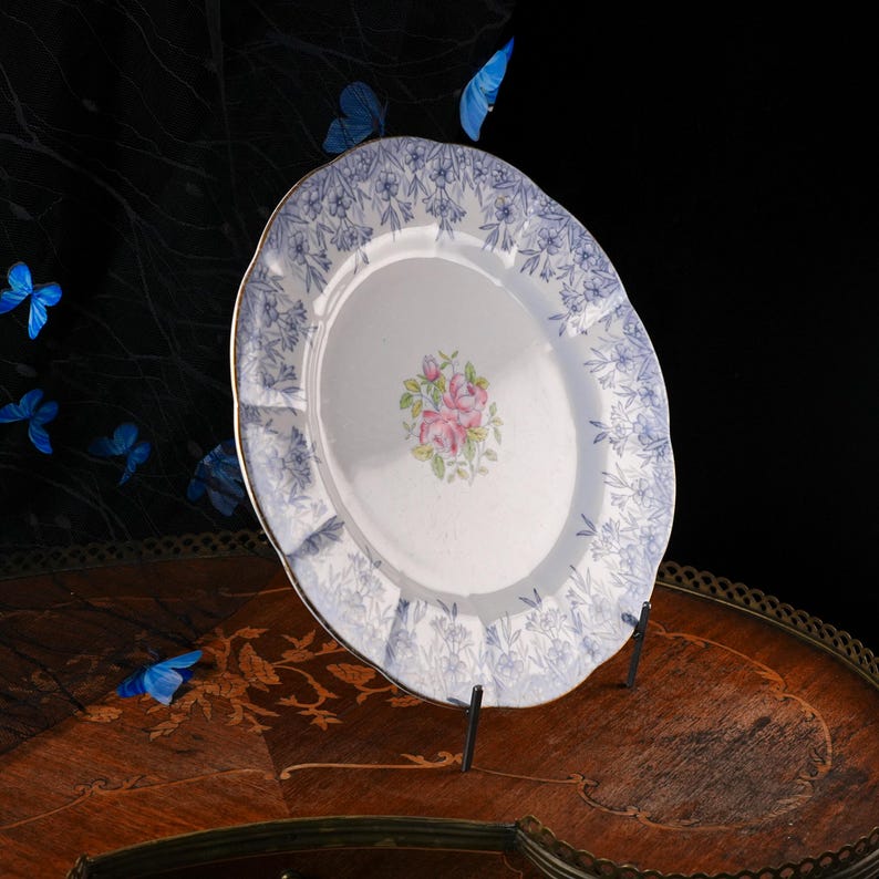 Puede incluir: Un plato redondo de cer&aacute;mica blanca con borde festoneado, decorado con un patr&oacute;n floral azul en el borde y un grupo central de rosas rosadas con hojas verdes. El plato se exhibe en posici&oacute;n vertical sobre un soporte de metal negro.
